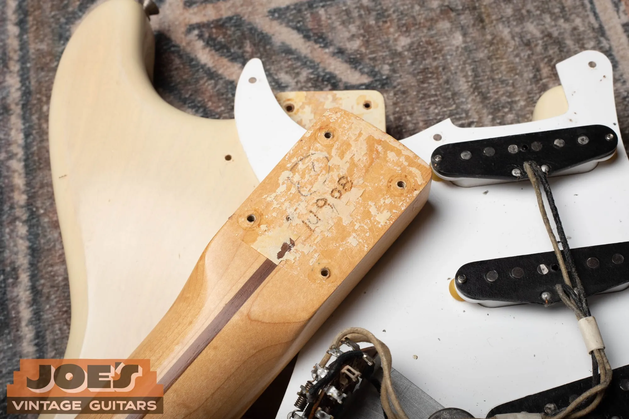 Close-up of the back of the neck heel of a 1956 Fender Stratocaster with the neck removed from the blonde body beside it, showing a serial number burned into the maple heel with a soldering iron along with a circled factory mark, with the pickguard assembly lying nearby.