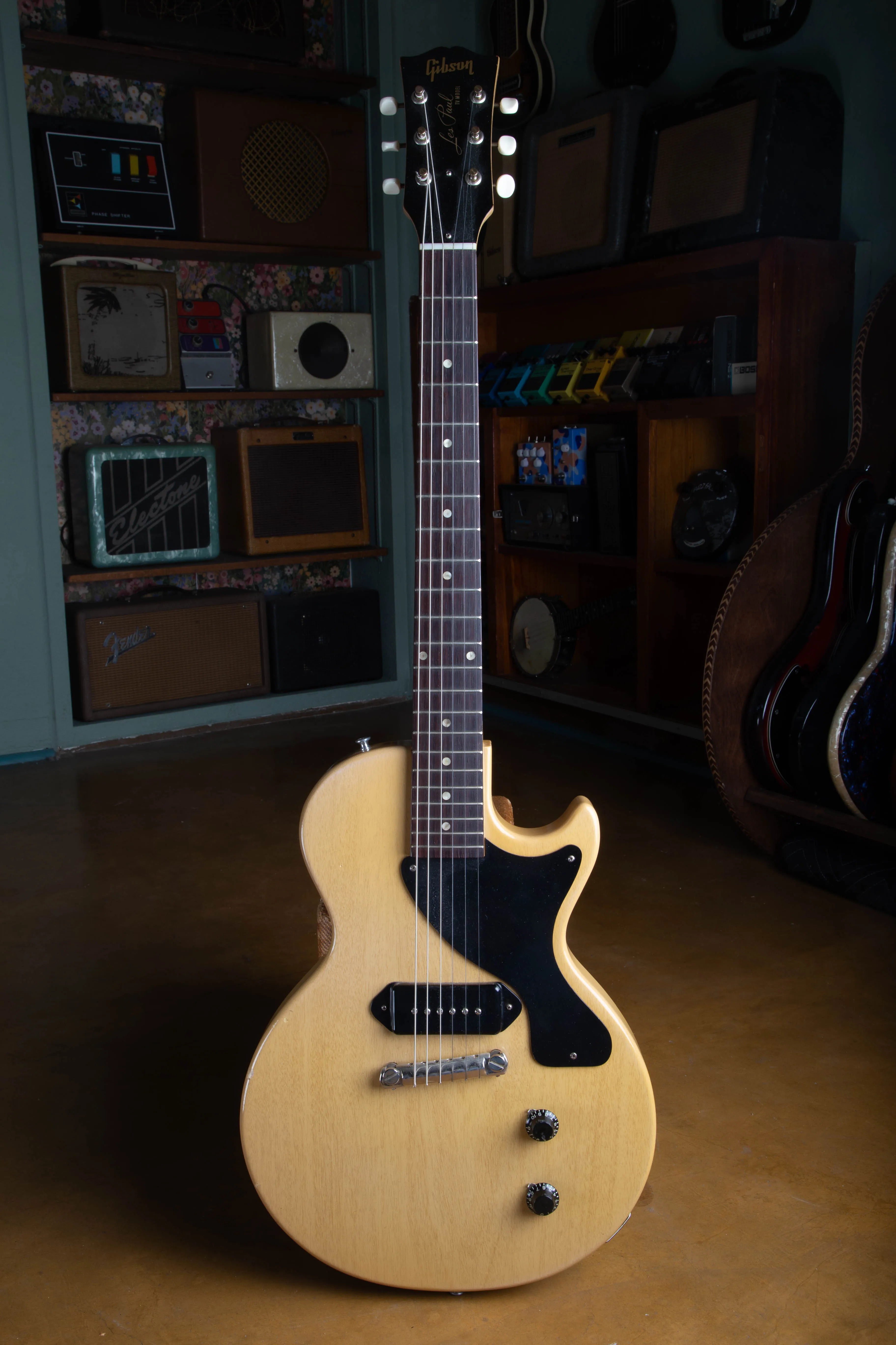Full front view of a 1957 Gibson Les Paul TV Model standing in a vintage guitar shop, showing the single-cutaway slab mahogany body in pale TV Yellow limed mahogany finish, single black dog-ear P-90 pickup, single-ply black pickguard, one-piece wraparound bridge, two black bonnet knobs, rosewood fingerboard with dot inlays, and a black headstock with gold Gibson and Les Paul TV Model silk-screen.