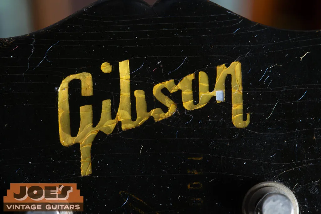 Macro close-up of a 1957 Gibson Les Paul headstock showing a Mother of Pearl “Gibson” logo with significant finish checking above it and a faded gold “Les Paul Model” silkscreen.
