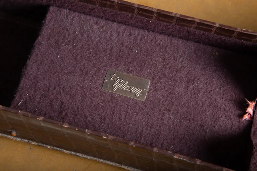 Detailed macro photograph of the original bronze-colored Gibson logo plaque located on the interior neck rest of an alligator chipboard case. The small, rectangular metal plate features the “Gibson” script in relief accompanied by a distinct five-pointed star icon, showing a natural aged patina and minor oxidation consistent with a 1950s accessory.