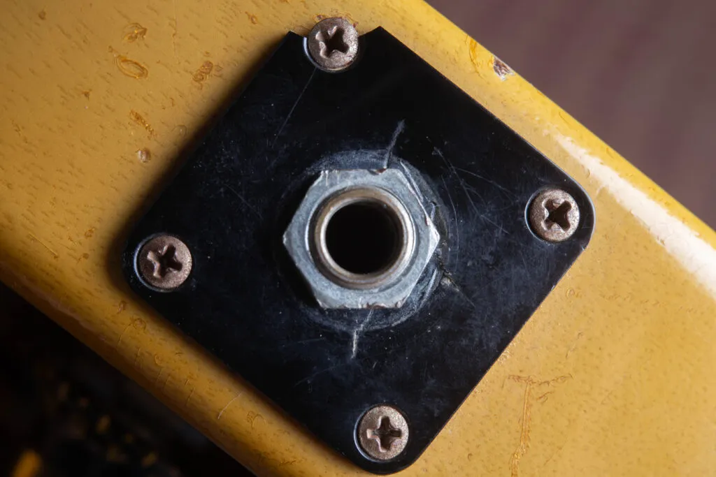 Detailed perspective of the side-mounted output jack on a vintage Gibson Les Paul Special. The image focuses on the original black plastic square jack plate, secured by four nickel-plated screws. The surrounding TV Yellow “limed mahogany” finish shows authentic nitrocellulose weather checking and natural patina around the plate edges.