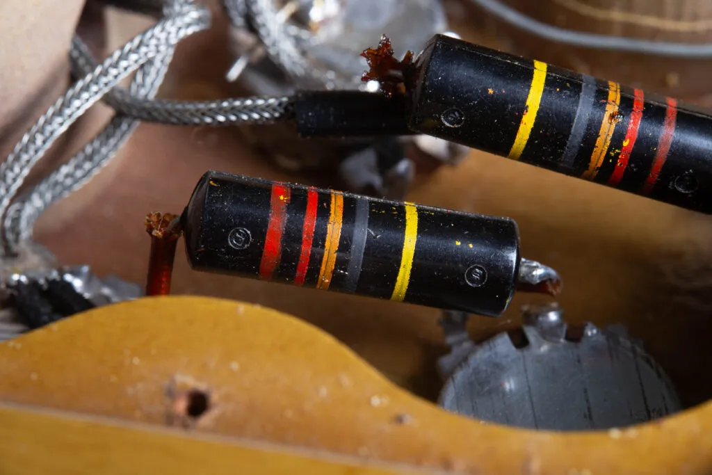 Close-up of two original “Bumblebee” capacitors inside a vintage Gibson Les Paul Special control cavity. The capacitors feature a black cylindrical body with distinctive colorful stripes (indicating capacitance and voltage) and are soldered to Centralab (CRL) potentiometers. The surrounding mahogany wood shows original factory routing, a rubber grommet for wiring, and authentic 1950s solder joints.
