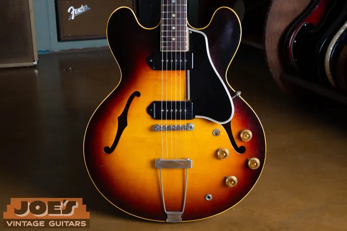Front of a sunburst 1959 Gibson ES-330 showing its two black dogear P-90 pickups, black pickguard, dot inlay rosewood fingerboard, gold bonnet knobs, and trapeze tailpiece, photographed in front of a vintage Fender amplifier.