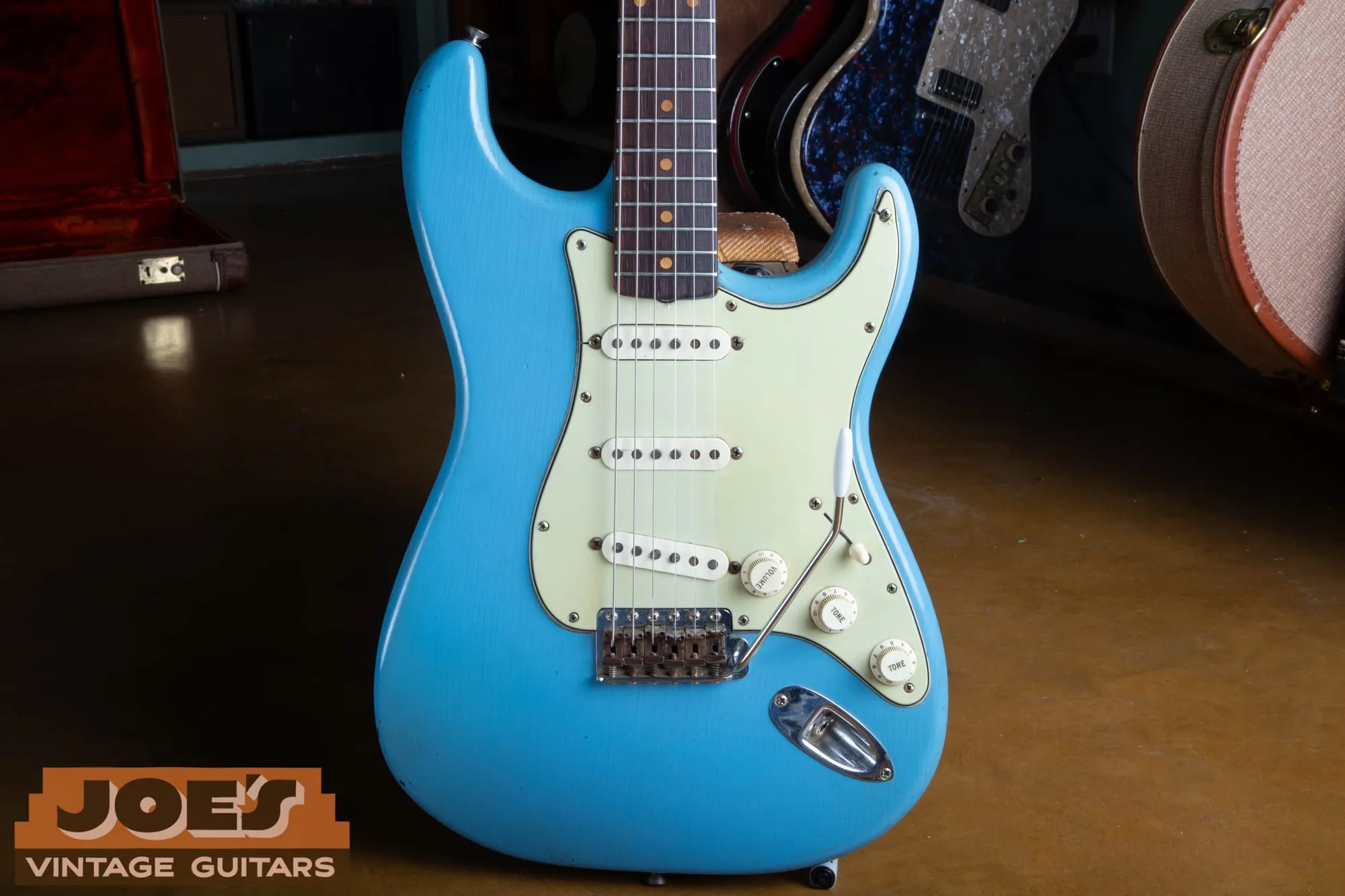 Front of a refinished 1960 Fender Stratocaster in a bright light blue that does not match any Fender custom color, with a mint pickguard, in a shop with other vintage guitars and cases behind it.