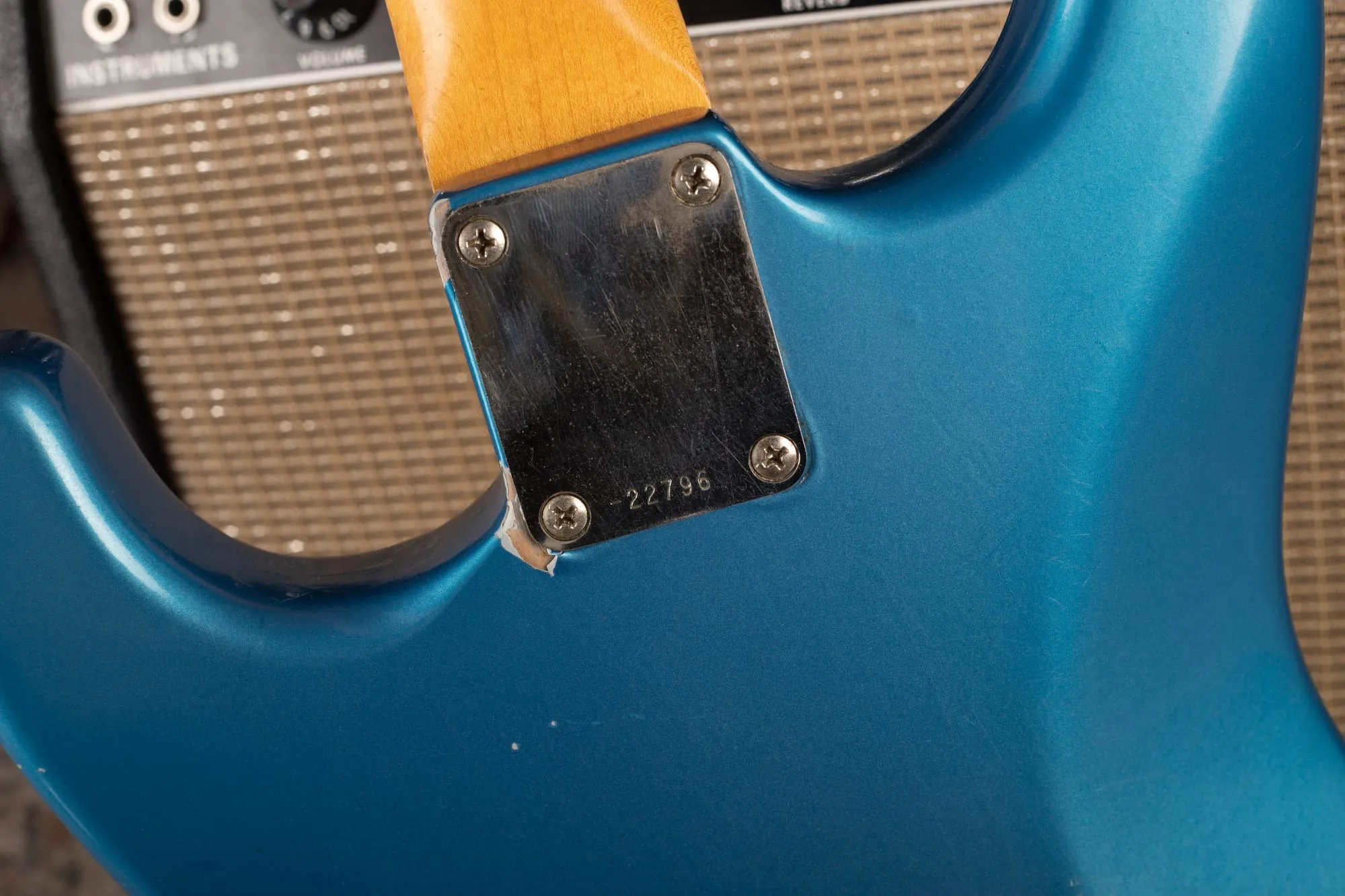 Back of a refinished 1960s Fender Stratocaster in blue metallic showing the serial number neck plate with finish bulging and ridging around the plate’s outline, odd rough texture, and a chip revealing grey primer, in front of a Fender amplifier.