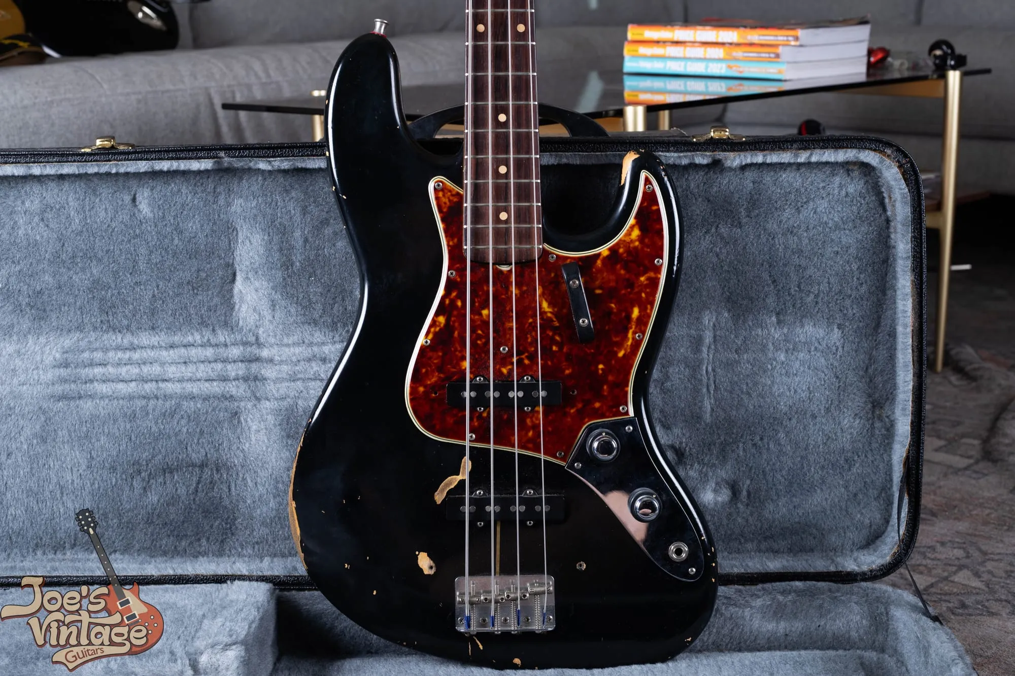 A refinished 1961 Fender Jazz Bass stack knob in black with a tortoiseshell pickguard, standing in a grey-lined case, with heavy wear showing bare wood directly beneath the black paint and no undercoat layer.