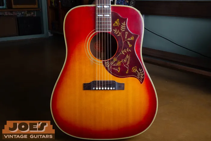 The cherry sunburst top of a 1961 Gibson Hummingbird showing the engraved pickguard with a hummingbird, trumpet flowers, and butterfly, and the rectangular rosewood bridge.