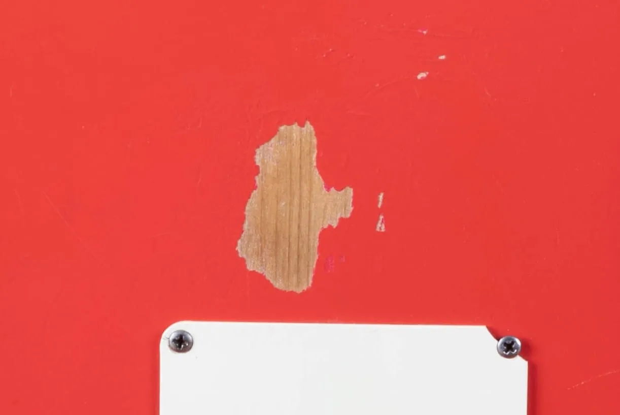 Close-up of the back of a 1964 Fender Stratocaster in Fiesta Red near the tremolo spring cover, with a chip in the red paint breaking directly to bare wood with no undercoat or sealer layer visible.