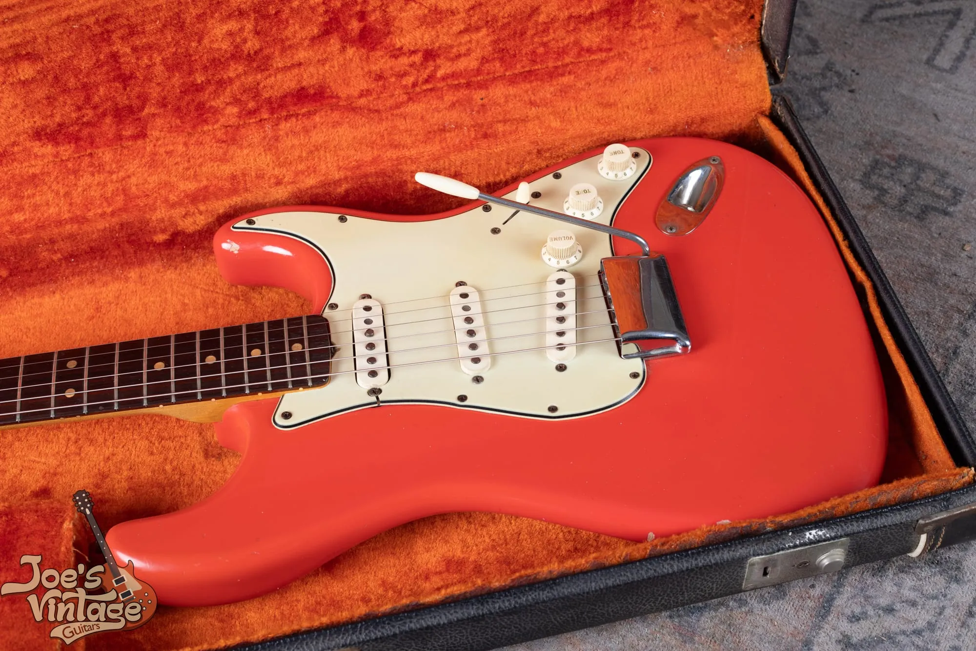 A 1964 Fender Stratocaster in Fiesta Red lying in its original black hardshell case with orange plush lining, showing the greenish mint three-ply pickguard, white plastic knobs and pickup covers, and rosewood fingerboard.