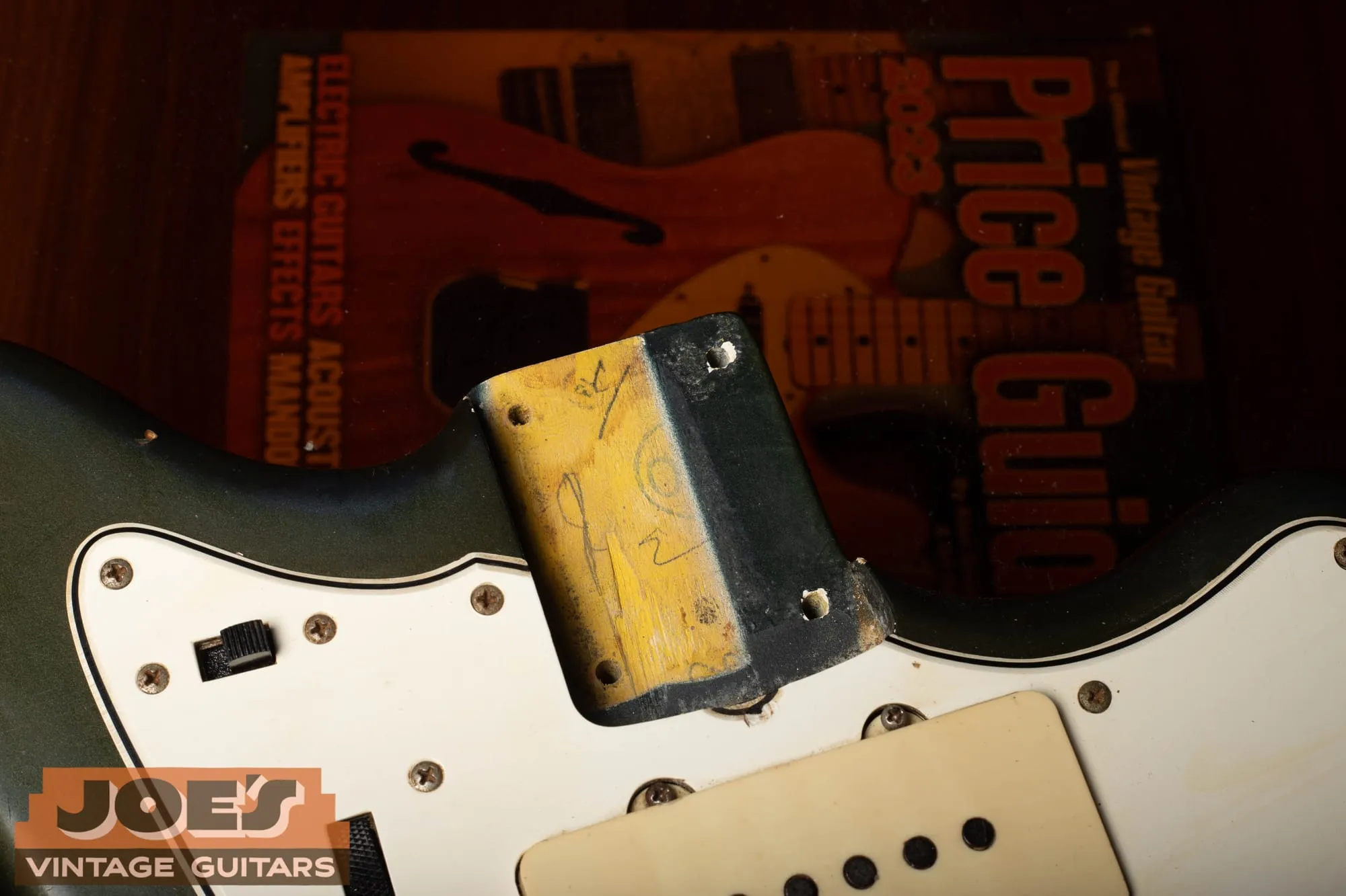 Close-up of the neck pocket of a 1965 Fender Jazzmaster in Charcoal Frost with the neck removed, showing a masked stripe of bare yellow sealed wood down the pocket with factory pencil markings, dark color coats surrounding it, and a Vintage Guitar Price Guide visible in the background.