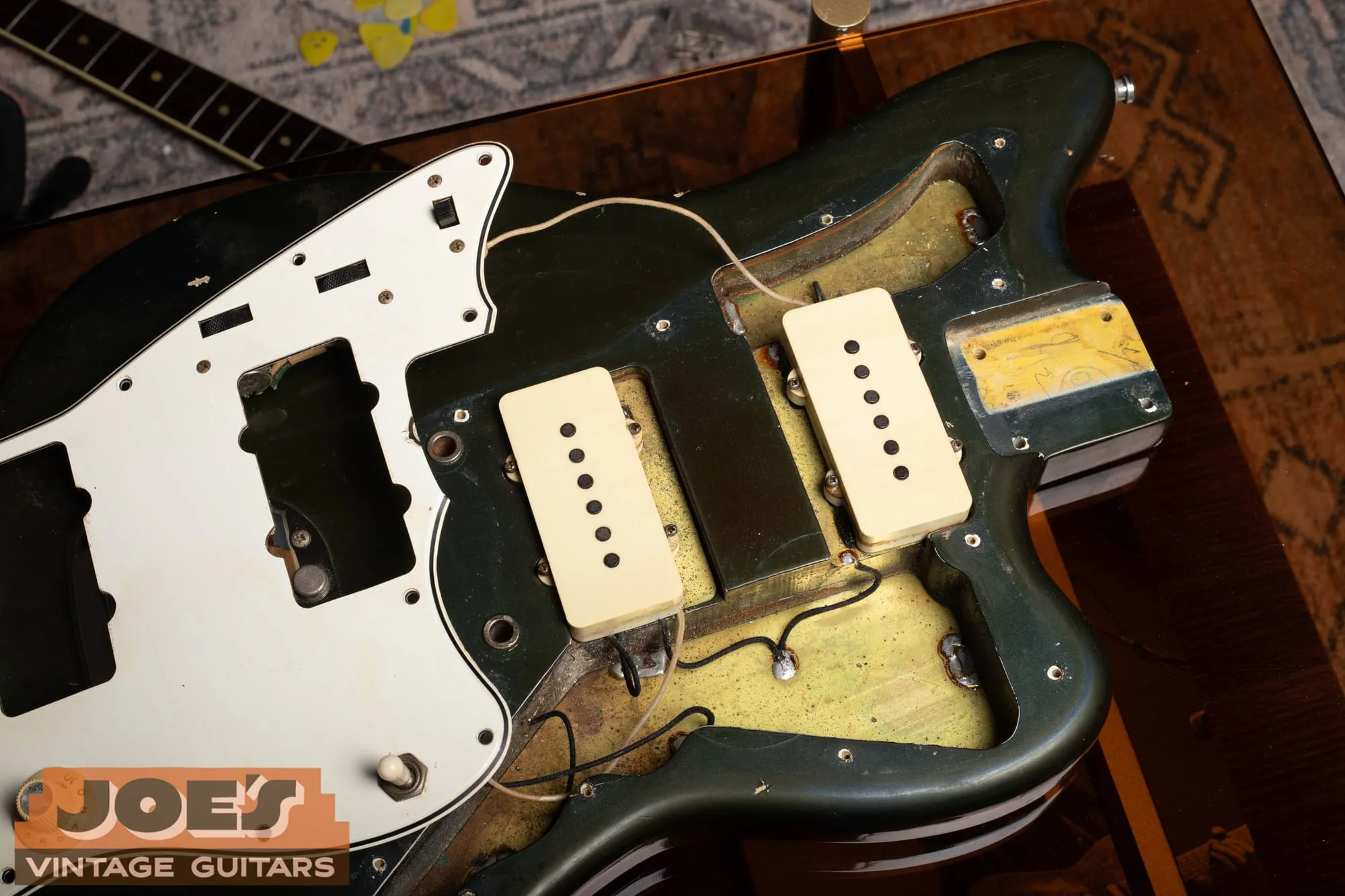 Under the lifted pickguard of a 1965 Fender Jazzmaster in Charcoal Frost, showing the original pickups sitting in their routes surrounded by yellow sealed wood where the color coats never reached, with the charcoal metallic visible at the body edges.