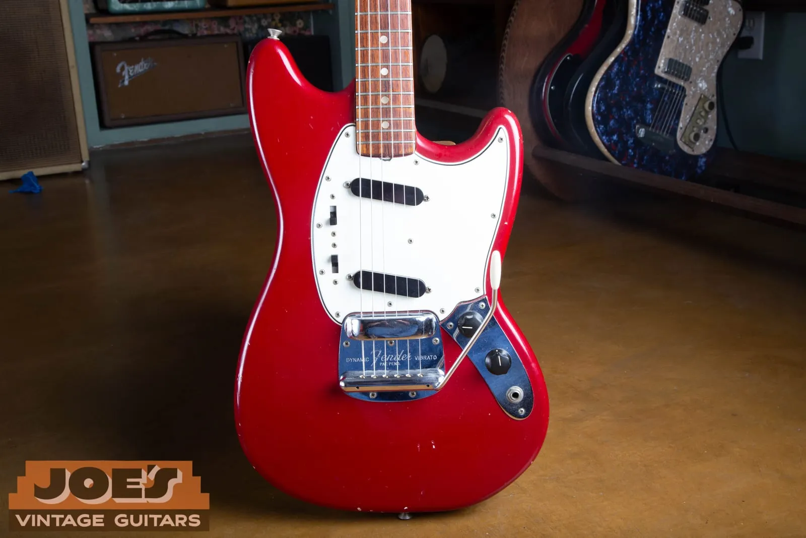 Front of a 1965 Fender Mustang in Dakota Red showing two angled single-coil pickups, two three-position slide switches, a white pearloid pickguard, and the chrome Dynamic Vibrato.