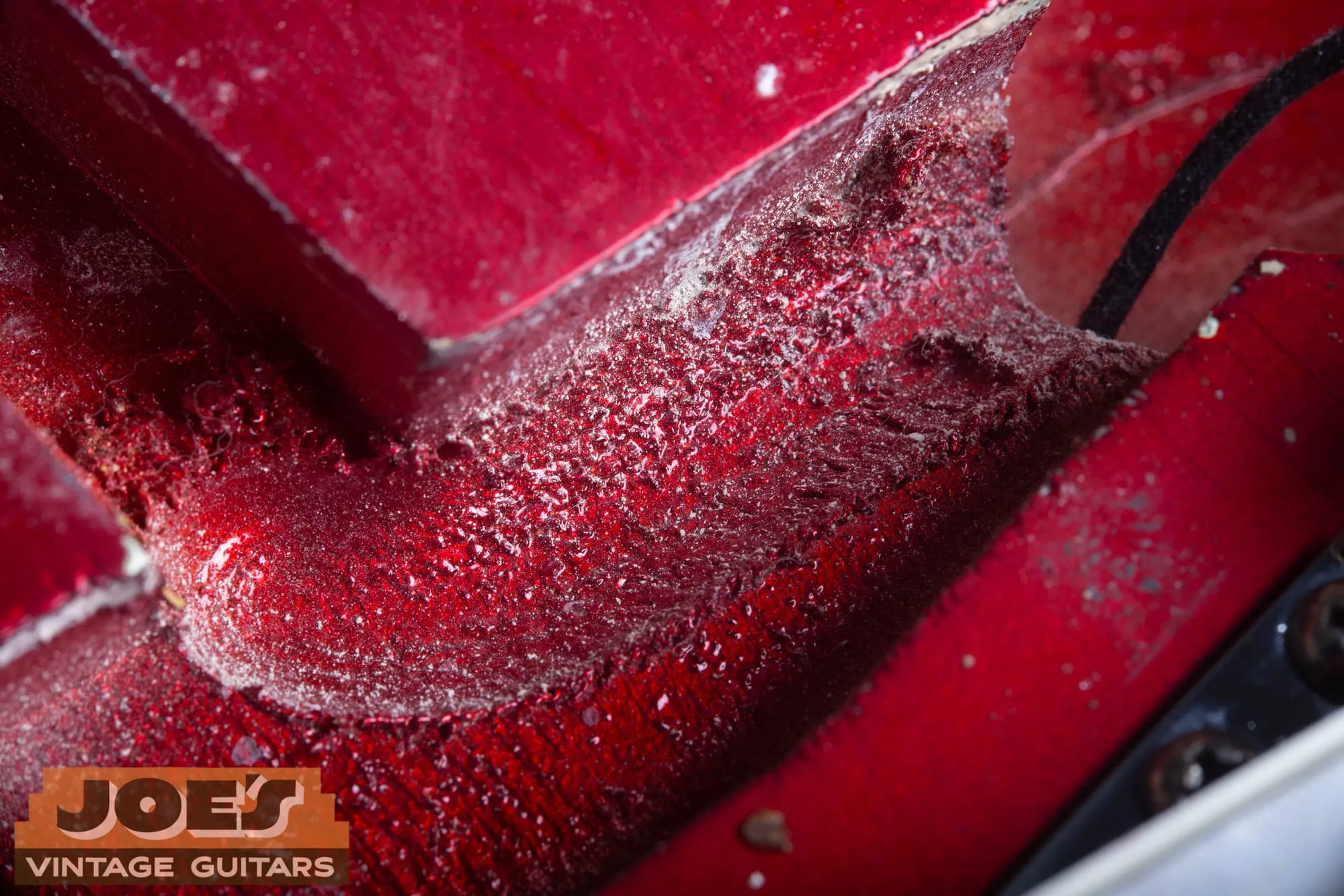 Extreme close-up of the bridge pickup route on a 1965 Fender Stratocaster in Candy Apple Red, showing a rough, torn worm route channel at the route edge with wood debris and fuzz sealed under the red paint.