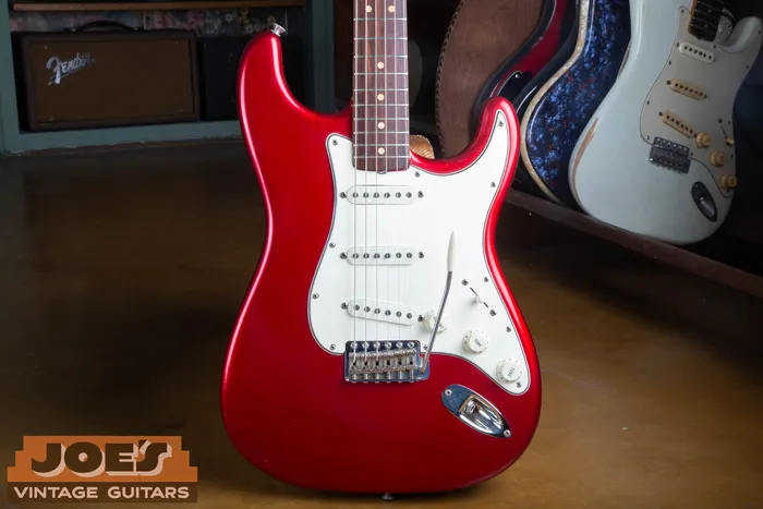 Front of a 1965 Fender Stratocaster in Candy Apple Red Metallic with a white three-ply pickguard, standing in front of a blackface Fender amplifier and other vintage Fender guitars, including a Sonic Blue Stratocaster.