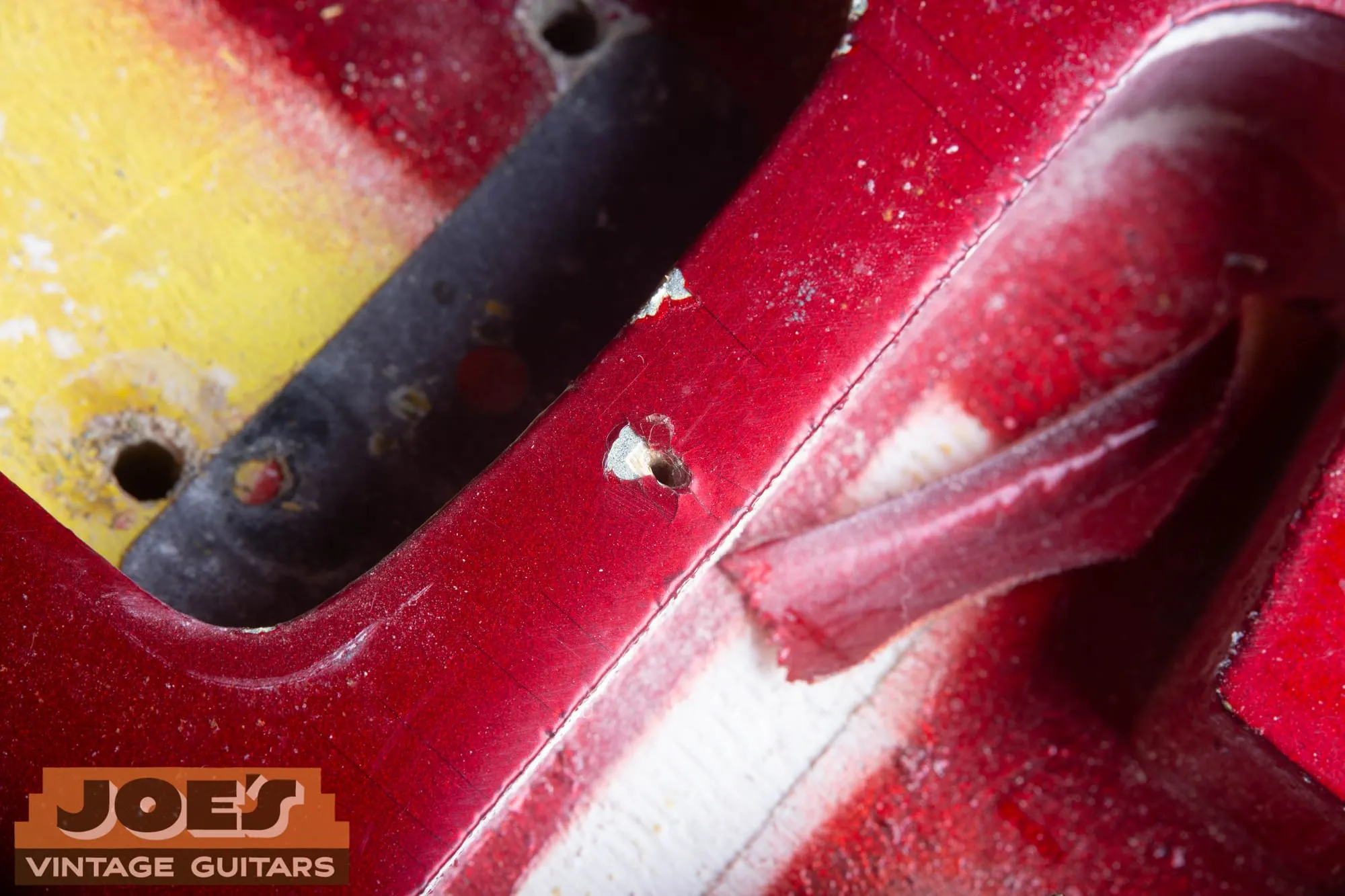 Extreme close-up under the pickguard of a 1965 Fender Stratocaster in Candy Apple Red showing a small factory nail hole with a chip at its rim revealing silver undercoat, a faint scribed arc in the finish, and the edge of a pickup route with yellow and dark sunburst-era color visible inside.
