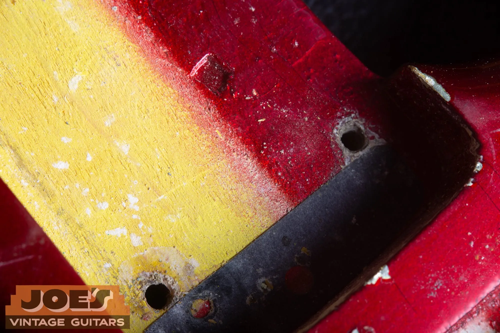 Close-up of the neck pocket area of a 1965 Fender Stratocaster in Candy Apple Red, showing the yellow-sealed pocket floor with red overspray feathering across it and heavy dust and shadow at the pocket edge.