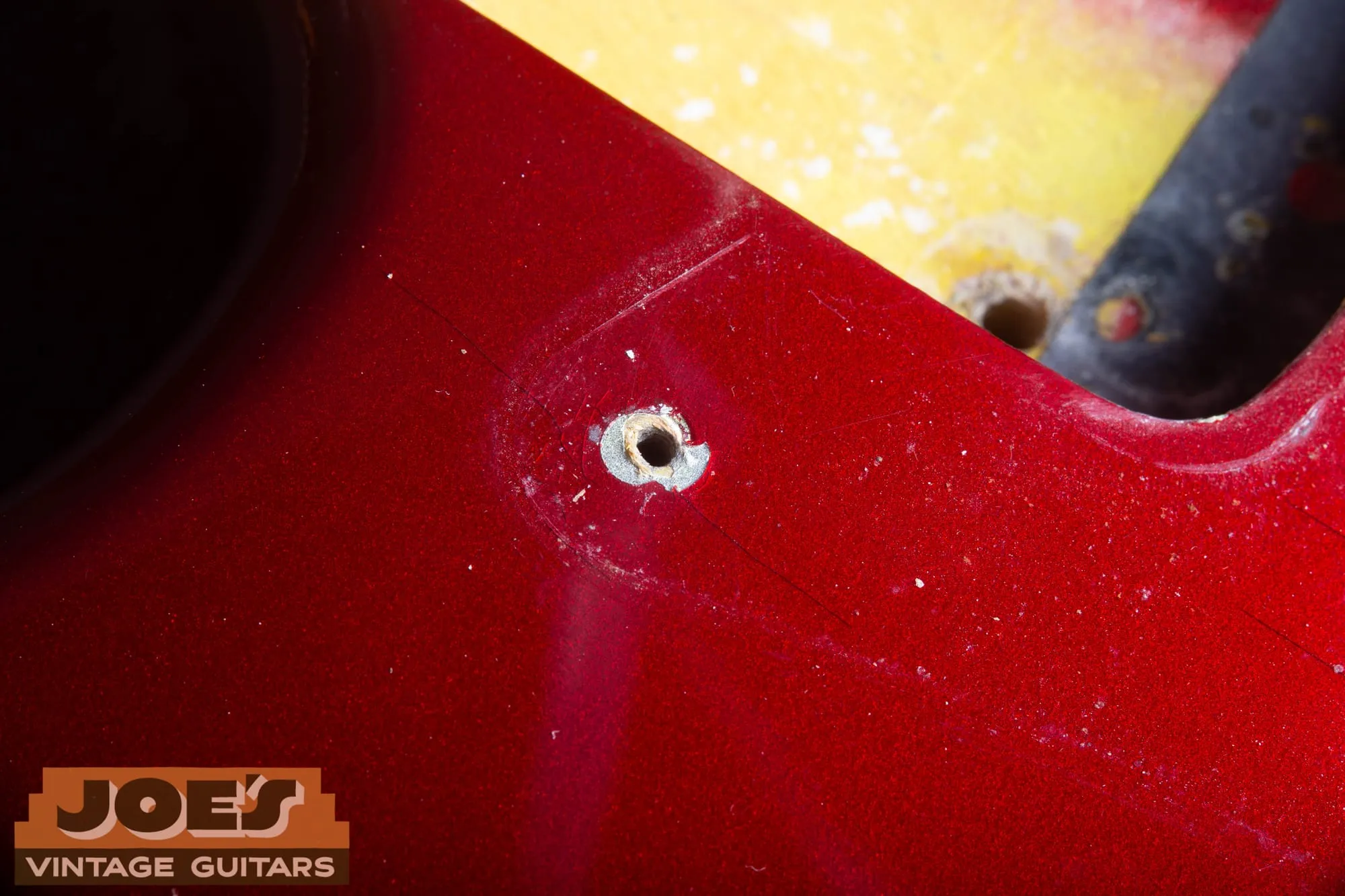 Extreme close-up of a pickguard screw hole on a 1965 Fender Stratocaster in Candy Apple Red, with a chip at the hole revealing the silver undercoat ring beneath the red and a pickup route with yellow sealed wood visible at the upper right.