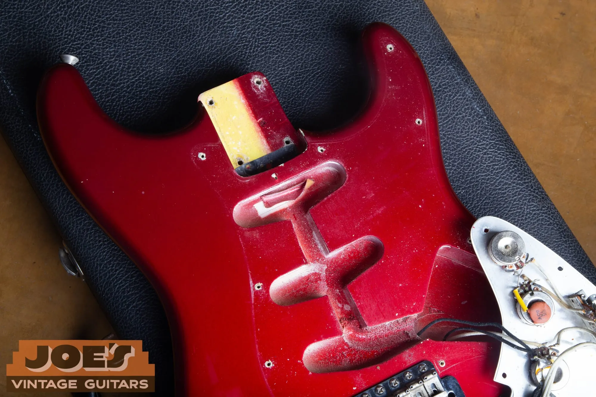 Top of a 1965 Fender Stratocaster body in Candy Apple Red with the pickguard removed and the loaded guard folded to the side, showing full red coverage in the pickguard footprint, dusty overspray in the pickup routes, and yellow showing through in the neck pocket.