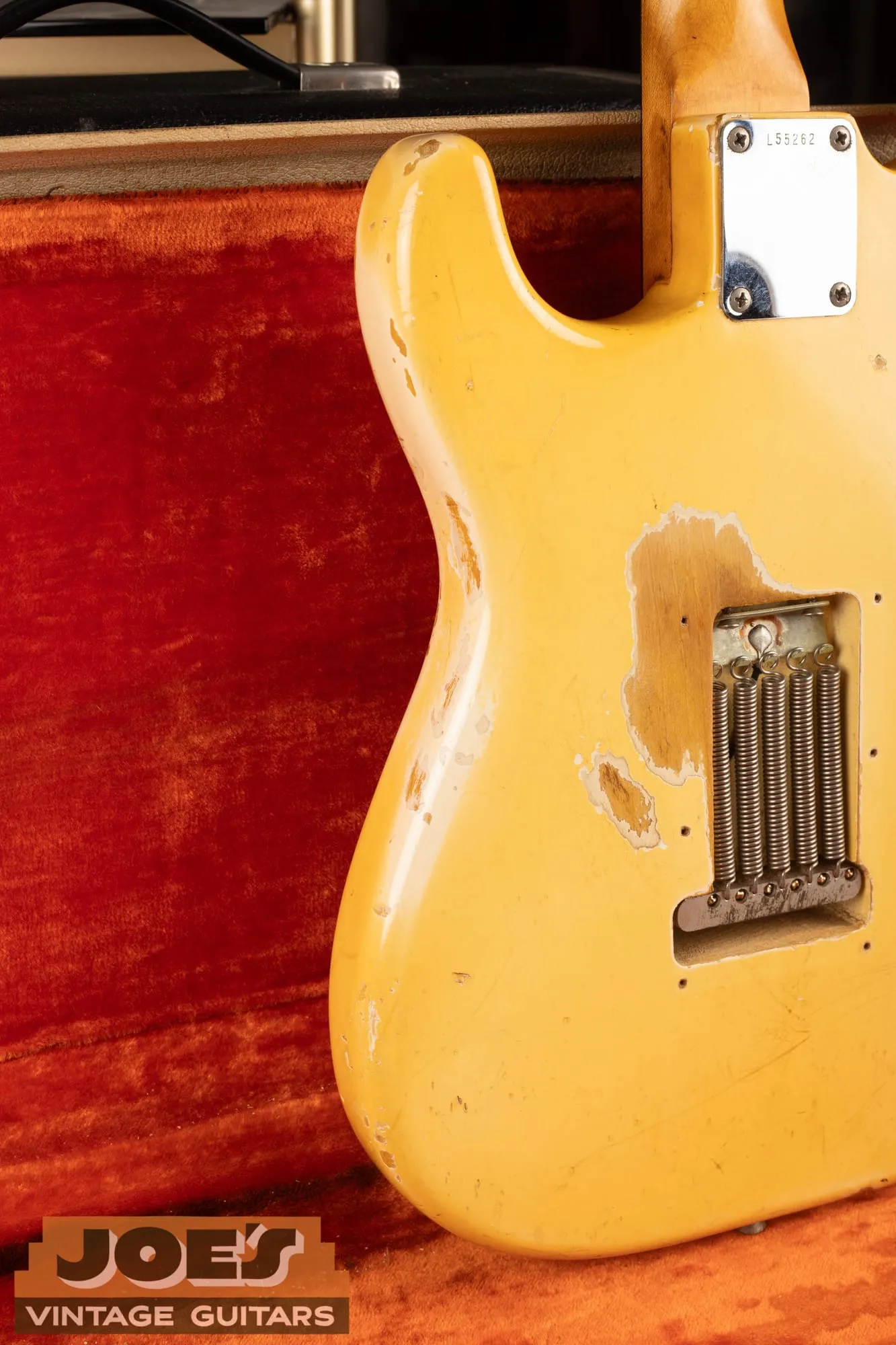 Back of a 1965 Fender Stratocaster in Olympic White in its case, with the trem cover removed showing the spring cavity, and large wear patches where the ambered cream top layer has worn through to reveal whiter paint beneath.