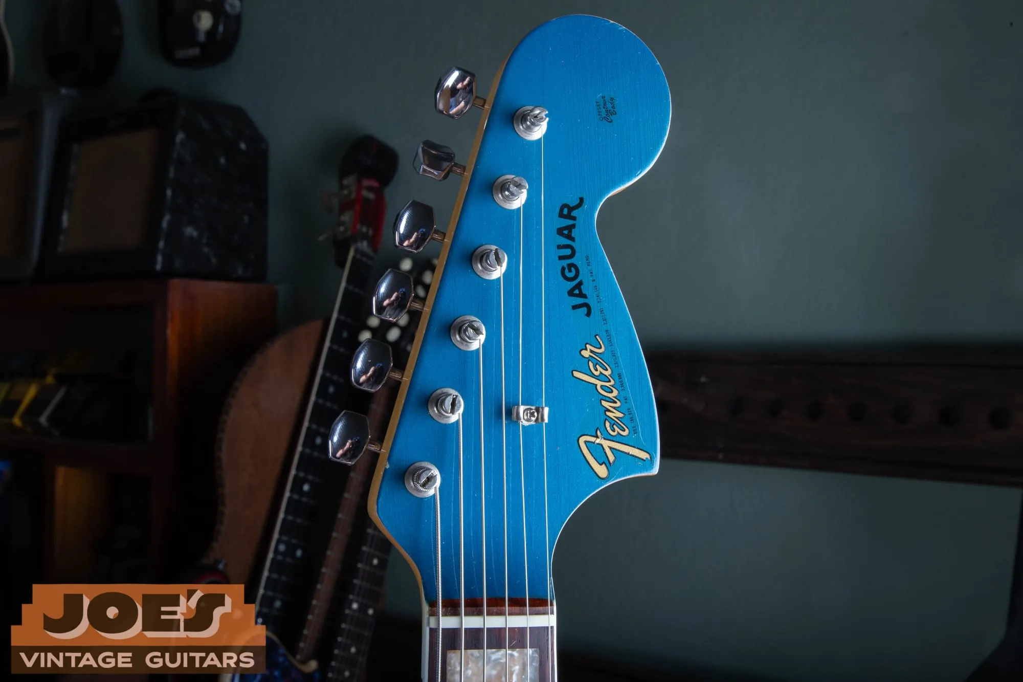 Matching painted headstock of a 1966 Fender Jaguar in Lake Placid Blue, showing the gold transition logo reading Fender Jaguar with patent numbers, single string tree, and Kluson tuners, photographed in front of a wall of vintage amplifiers.