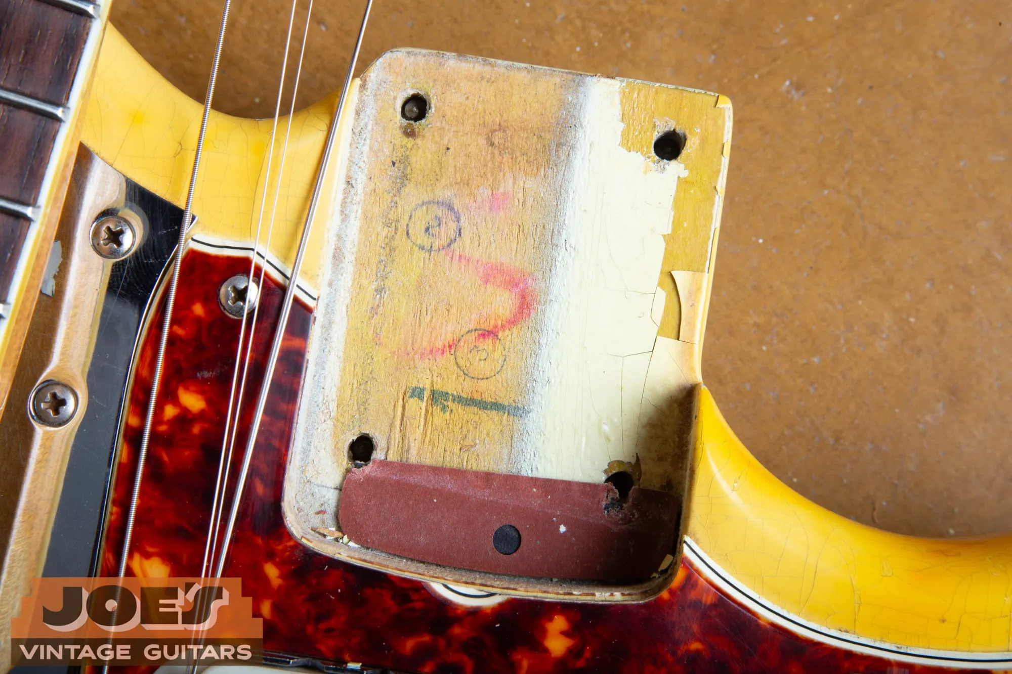 Neck pocket of a 1966 Fender Jaguar in Olympic White with heavily checked and ambered finish, the pocket floor showing bare wood with red and black factory crayon markings and circled numbers, a whiter band of paint at one edge, and a rust-red fiber shim at the pocket end, with a tortoiseshell pickguard around it.