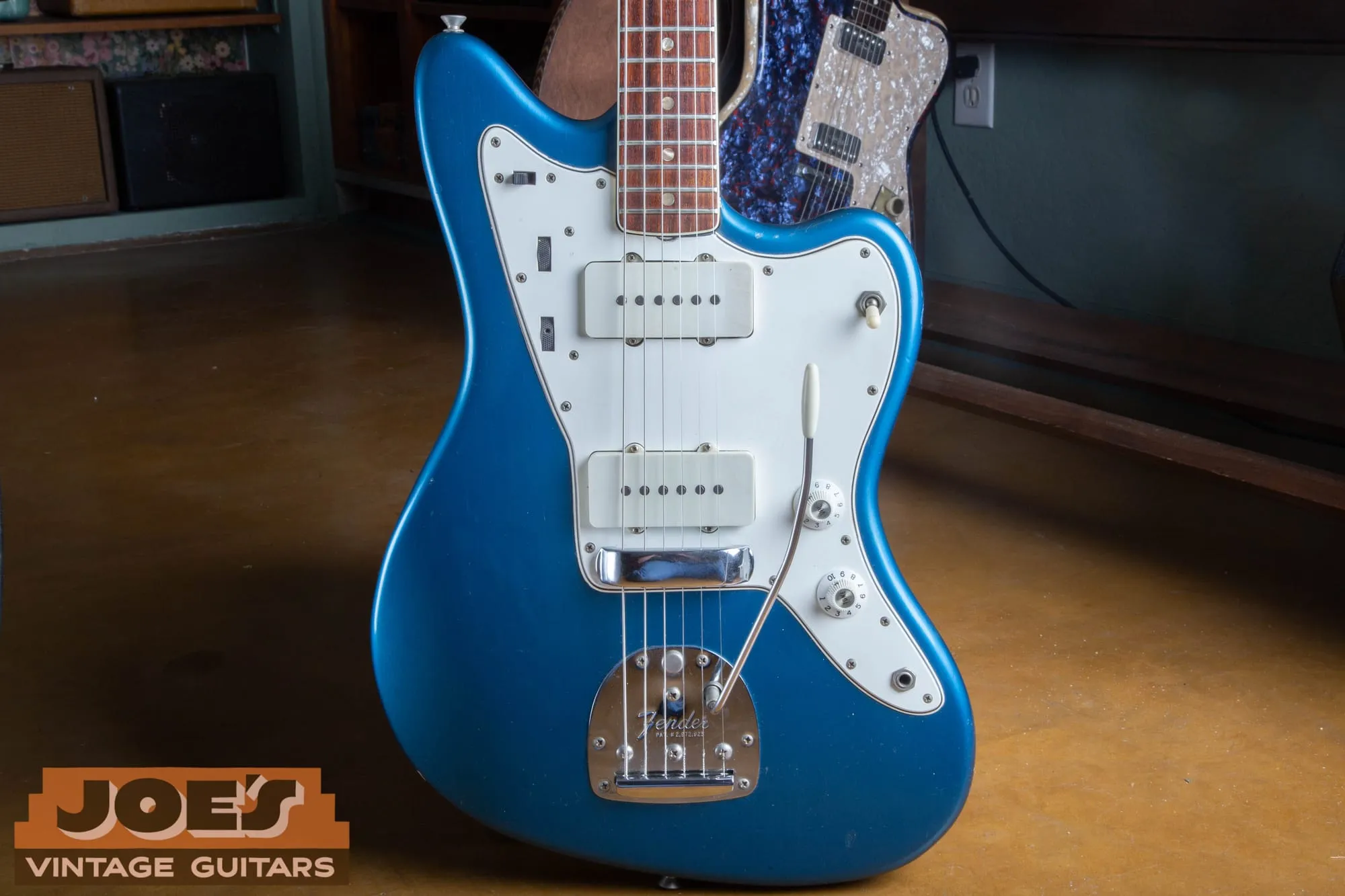 Front of a 1966 Fender Jazzmaster in Lake Placid Blue with a white pickguard, photographed in a vintage guitar shop with a blue sparkle guitar visible behind it.