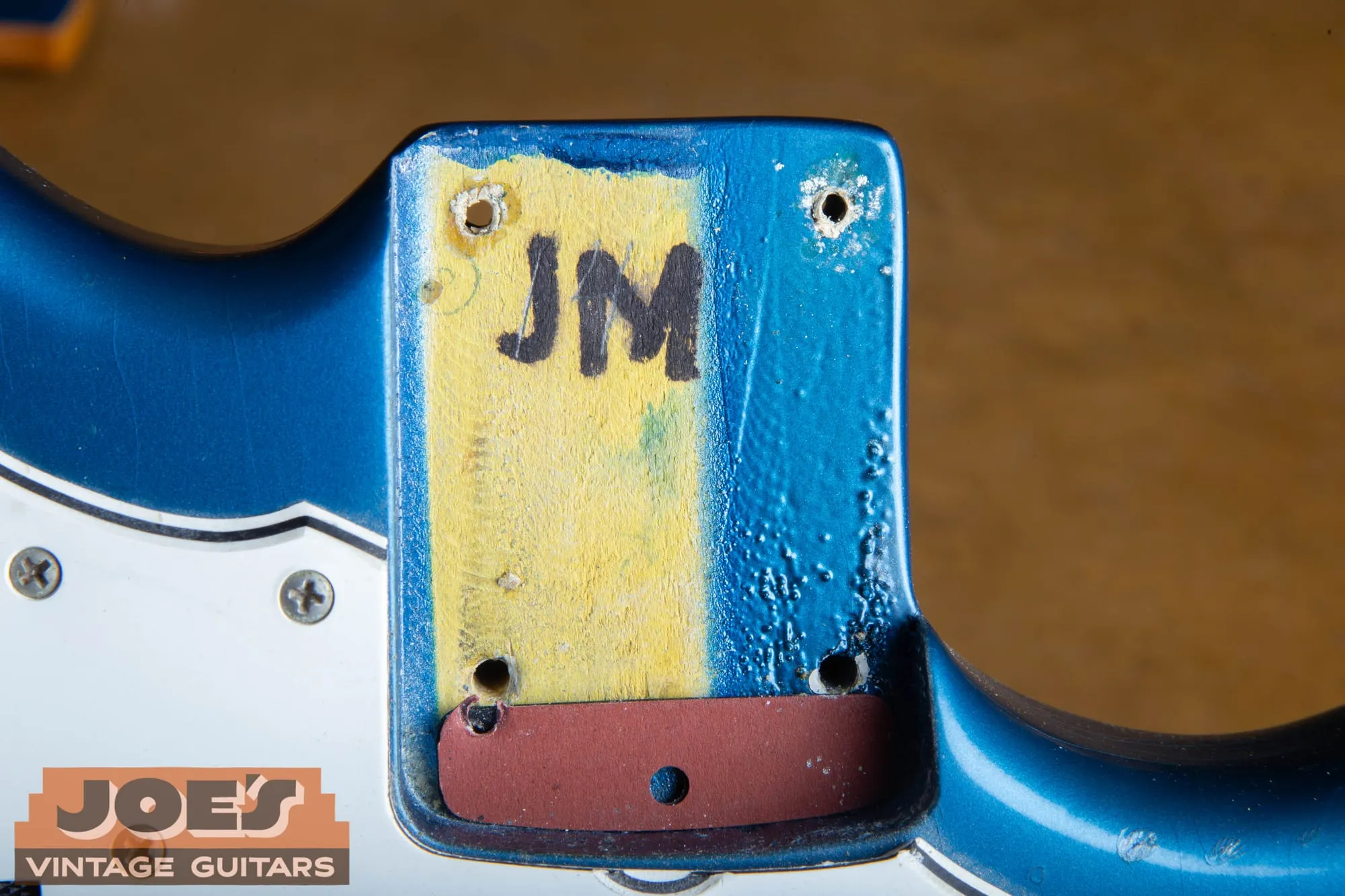 Neck pocket of a 1966 Fender Jazzmaster in Lake Placid Blue with the neck removed, showing a masked yellow stripe down the center of the pocket with JM written in black marker, blue overspray on either side, bubbly finish texture at the pocket edge, and a rust-red fiberboard factory shim at the end of the pocket.