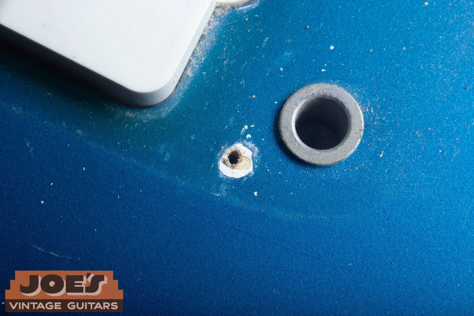 Extreme close-up of a pickguard screw hole on a 1966 Fender Jazzmaster in Lake Placid Blue next to a bridge post thimble, with the chipped edge of the screw hole revealing a bright white undercoat beneath the blue.