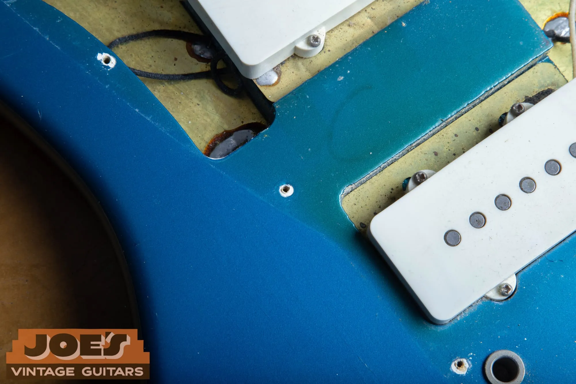 Under the pickguard of a 1966 Fender Jazzmaster in Lake Placid Blue, showing bright blue in the exposed areas transitioning to thin dusty coverage and large patches of exposed yellow-stained sealed wood around the pickup routes, with original wiring visible.