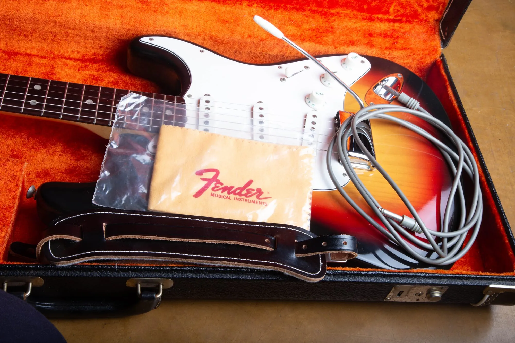 Open black Fender case with orange plush lining holding a 1967 Stratocaster, with a Fender Musical Instruments polish cloth, a coiled vintage cable, a leather strap, and the tremolo arm as original case candy.
