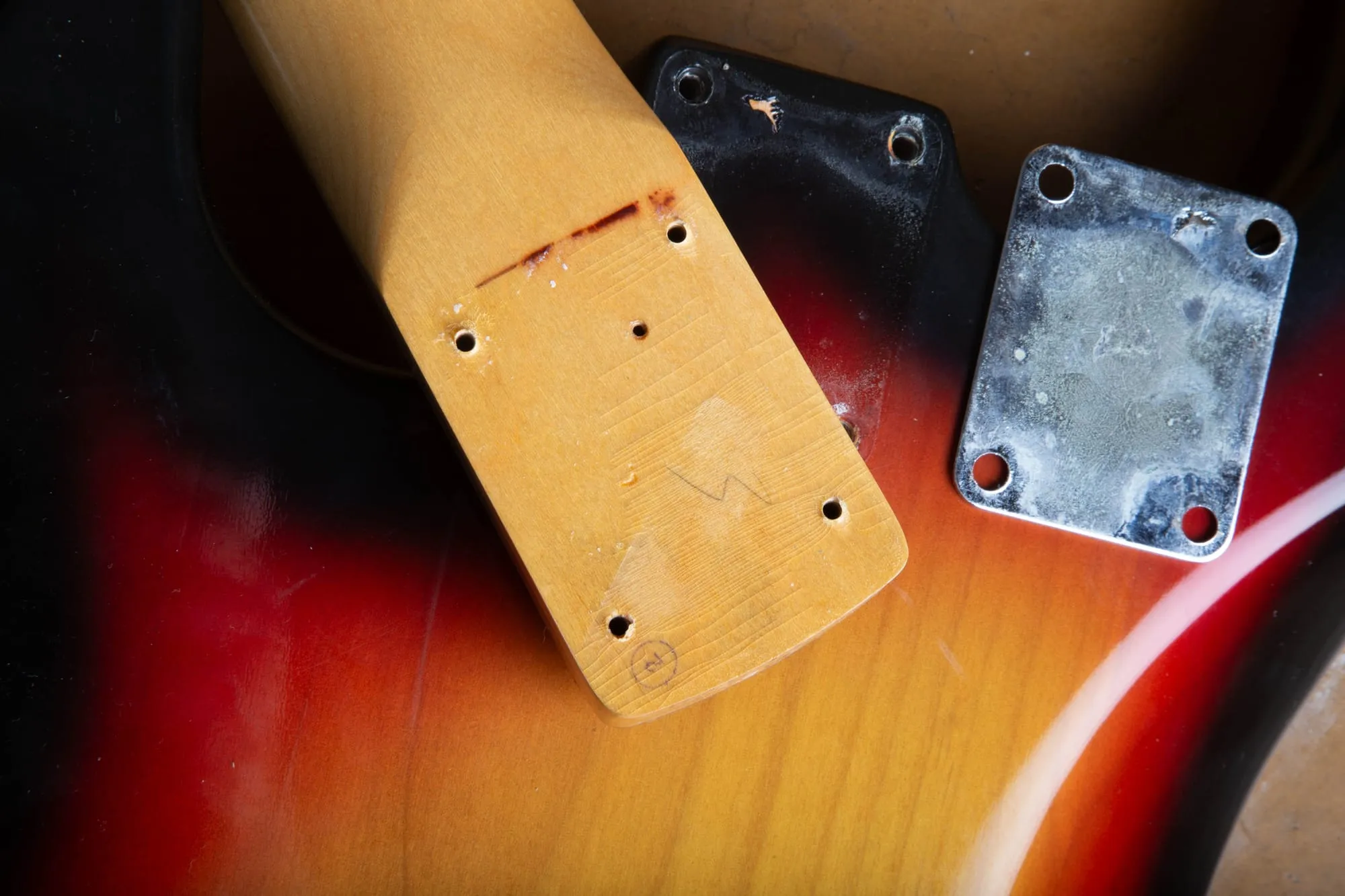 Underside of a 1967 Fender Stratocaster neck heel out of the body, showing a red paint stick crayon mark, pencil assembly marks, and a small ink stamp, next to the chrome F neck plate.