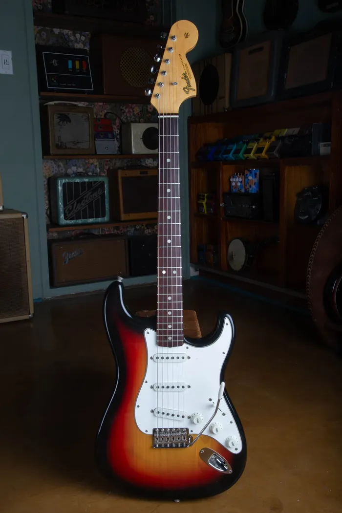 A 1967 Fender Stratocaster in three-tone sunburst standing in a vintage guitar shop, with a large CBS headstock, rosewood fingerboard, and aged white pickguard.