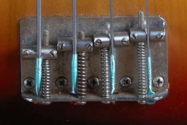The chrome bridge of a 1973 Fender Jazz Bass with four threaded saddles and the strings anchored through the bridge plate.