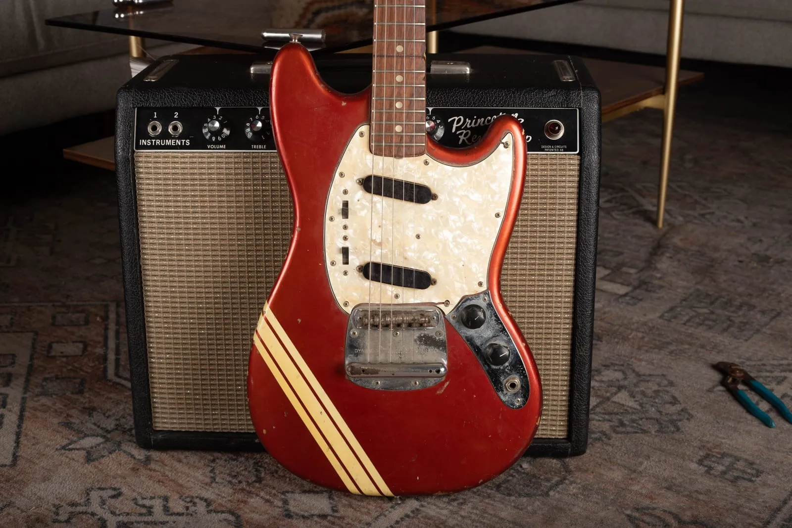 Front of a 1972 Fender Competition Mustang in Competition Red with a racing stripe and heavy honest player wear, next to a Fender Princeton amp.