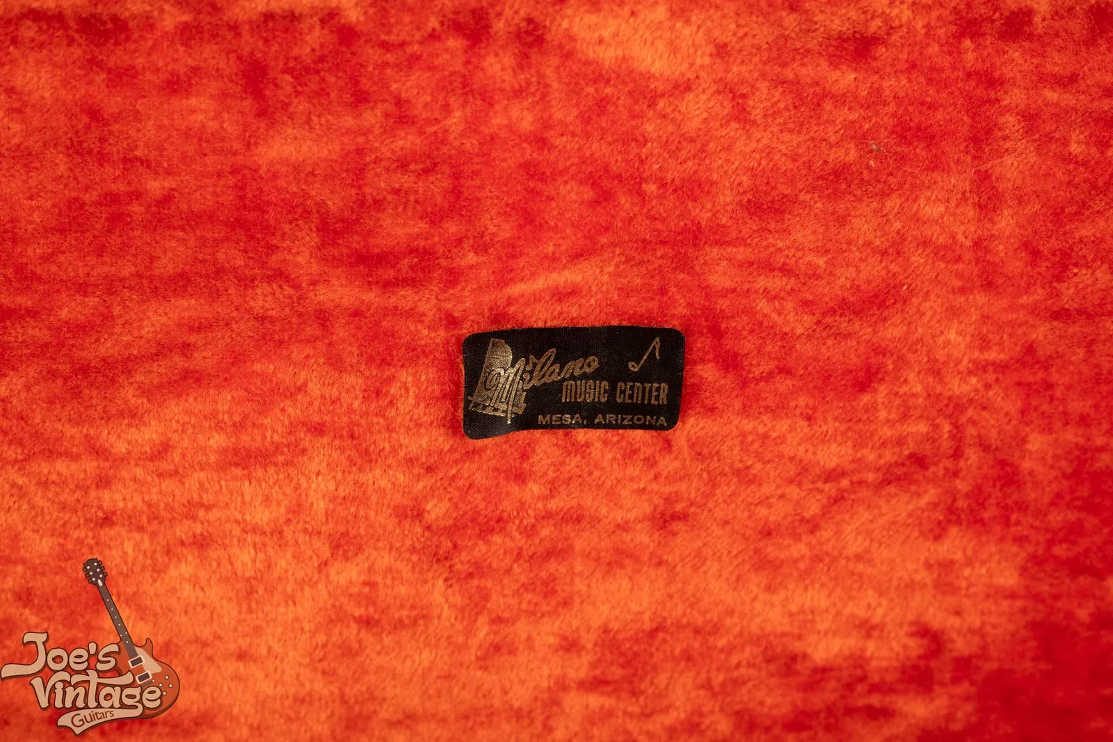 Interior of a vintage Fender hardshell guitar case with orange plush lining and a Milano Music Center, Mesa, Arizona dealer sticker.