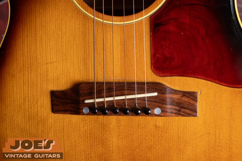 The rosewood bridge of a vintage Gibson flat-top guitar photographed from above, with a light colored saddle standing only slightly proud of the bridge surface and eight bridge pins in a row.
