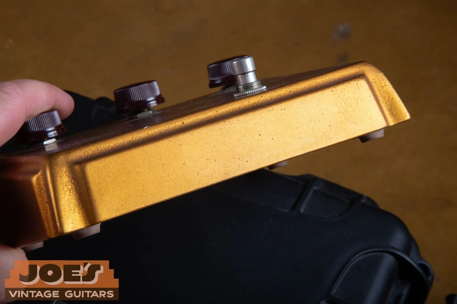 Side profile of a gold Klon Centaur held in a hand, showing the tall cast enclosure, the three knobs, and the footswitch on the sloped face.
